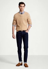 Beige cable-knit sweater layered over a blue striped shirt, paired with dark jeans and brown loafers. Simple design, textured knits and classic colors.