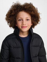 Enfant aux cheveux bouclés portant une doudoune noire sur un haut bleu marine, souriant doucement sur un fond clair uni.