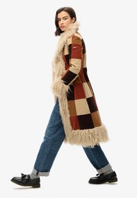 Abrigo de patchwork que presenta cuadrados en color marrón, beige y rojo, con un cuello y un dobladillo beige y peludo. Combinado con jeans azules y mocasines negros.