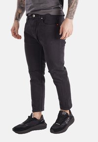 Jeans in denim nero con una vestibilità slim, cinque tasche e un evidente dettaglio del logo rosso. Abbinati a scarpe nere con accenti testurizzati.