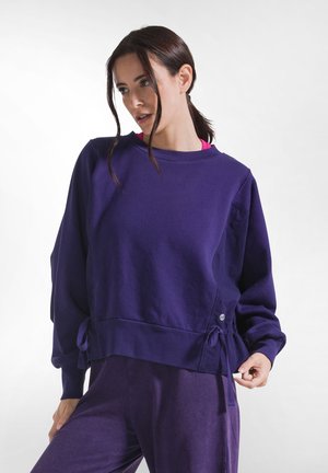 Vrouw met donker haar in een staart draagt een losse paarse trui met zijstrikjes en bijpassende broek, poserend tegen een effen witte achtergrond.