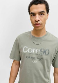 Zielona bawełniana koszulka z krótkim rękawem, z nadrukowanym tekstem "Core90" i "Creative Dept." w kontrastowych kolorach oraz teksturowanym wykończeniu.
