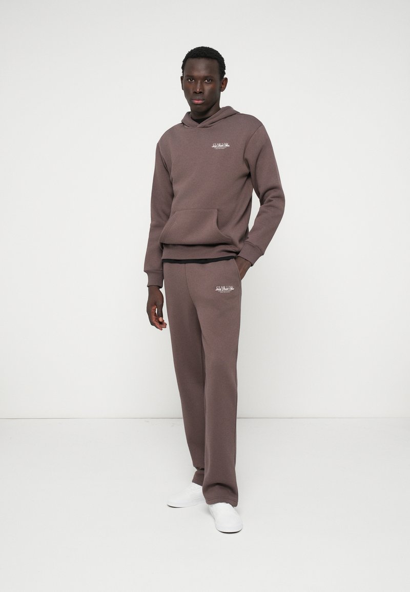 Brązowa bluza z kapturem i przednią kieszenią, zestawiona z dopasowanymi spodniami. Minimalistyczny design z haftowanym logo, na jasnym tle.