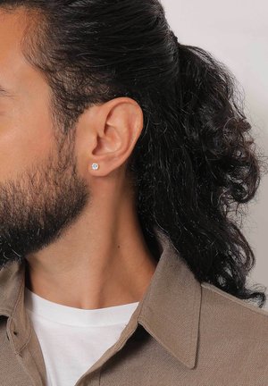 Silberne Stud-Ohrringe mit floralen Design. Nahaufnahme eines männlichen Modells mit dunklem, lockigem Haar, der ein beiges Hemd über einem weißen T-Shirt trägt.