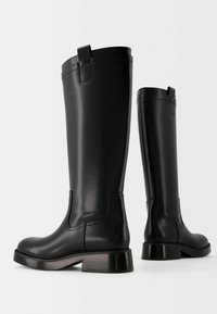 Par de botas negras de cuero altas con tacones bajos y tiradores sobre un fondo claro y liso.