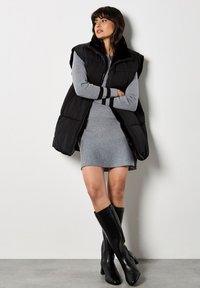 Schwarze Steppweste mit hohem Kragen, getragen über einem grauen Kleid. Die Weste verfügt über einen Reißverschluss und gestreifte Akzente an den Ärmeln. Schwarze kniehohe Stiefel.