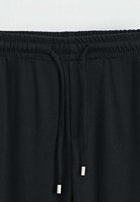 Un pantalon de jogging noir avec une taille élastique et un cordon de serrage avec des embouts métalliques, fabriqué à partir d'un tissu lisse et offrant une coupe décontractée.