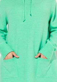 Mintgrön cardigan med huva, ribbade ärmslut och två framfickor. Mjuk textur och rak siluett med fullängdsdragkedja.