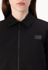 Musträpiga musta jope, millel on krae, esine tõmblukk ja tekstuuriga kangas. Vasakul küljel on väike must logopatch.
