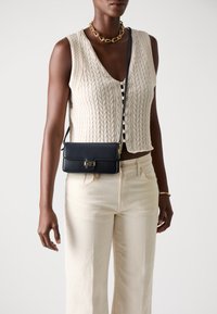 Svart lær crossover bag med gull hardware, rektangulær form og glatt tekstur, båret over en lys strikket vest og beige bukser.