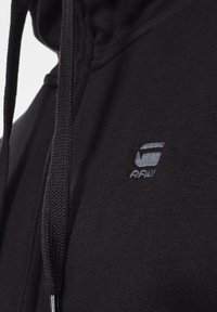 Felpa nera con tessuto testurizzato, dotata di un logo grigio chiaro. Include un cappuccio regolabile e una zip frontale. Design semplice.