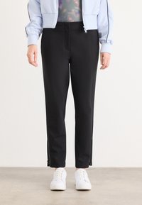 Zwarte, op maat gemaakte broek met zijritsen en een gladde textuur, gecombineerd met witte sneakers en een lichtblauwe bomberjack.