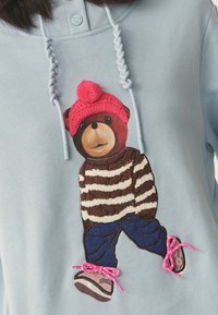 Kék kapucnis pulóver medve grafikával. A medve rózsaszín kötött sapkát, csíkos pulóvert és kék farmert visel, a cipőkön pedig rózsaszín cipőfűzők vannak.