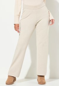 Onderlichaam van een persoon in beige gebreide broeken, een bijpassende trui en tan suede enkellaarsjes staan tegen een witte achtergrond.