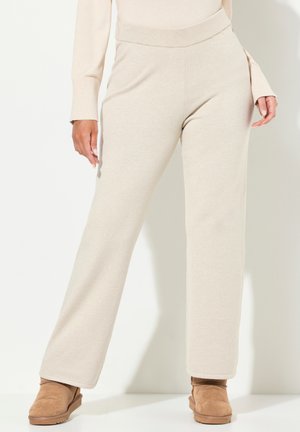 Underkroppen af en person iført beige strikbukser, matchende sweater og brune ruskinds ankelstøvler, stående foran en hvid baggrund.