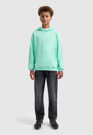 Tienerjongen staand, draagt een mintgroene hoodie, zwarte spijkerbroek en zwarte sneakers tegen een effen lichtgrijze achtergrond.