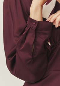 Bluză burgundy cu mâneci lungi, având detaliu cu buton la manșetă și material moale, fin. Designul plisat adaugă dimensiune.