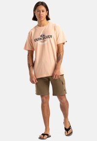 Camiseta de algodón melocotón con el logo de "QUIKSILVER", combinada con pantalones cortos verdes oliva que tienen bolsillos y un pequeño logo. Chanclas negras.