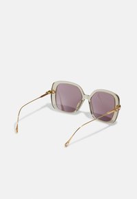 Gafas de sol cuadradas con montura clara y transparente, lentes púrpuras y acentos de metal dorado en las patillas. Textura de superficie suave en toda la pieza.