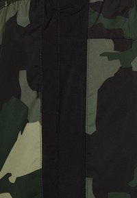 Zelené maskáčové cargo šortky s černou síťovinou. Mají elastický pas a boční kapsy. Tkanina vypadá hladce.