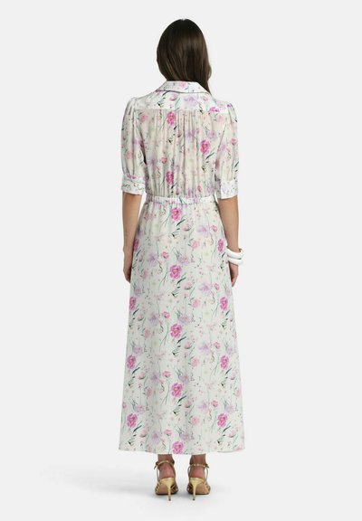 Femme portant une longue robe blanche à fleurs avec des manches bouffantes et un col, assortie à des chaussures à talons hauts beiges, vue de dos.