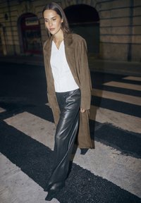 Manteau long en daim marron, chemise blanche à boutons, pantalon noir à motifs avec une texture brillante, marchant sur un passage piéton la nuit.