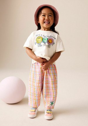 Lächendes junges Mädchen mit rosa Hut, weißem T-Shirt mit der Aufschrift "It's a Smiley World", bunten karierten Hosen und pastellfarbenen Turnschuhen steht neben einem pinken Ball.