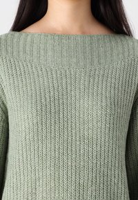 Pull en maille vert clair avec un col large côtelé et un corps texturé. Le tissu semble doux et confortable avec une silhouette ample.