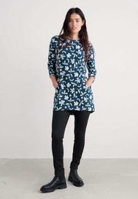 Ženska stoji z rokami v žepih, oblečena v teal dolgi rokav cvetlični tunik, črne oprijete hlače in črne gležnjarje, pred enobarvno ozadjem.