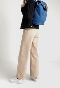 Zaino in tessuto blu con dettagli in pelle marrone, indossato sopra una giacca nera. La persona indossa pantaloni beige a gamba larga e sneakers bianche.