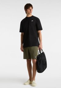 T-shirt nera in cotone, pantaloni corti verde oliva, zoccoli gialli, mentre tiene una borsa nera. Design semplice, maniche corte, logo discreto sulla t-shirt.