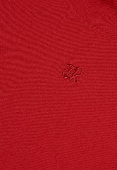 Suéter de punto rojo con cuello redondo y un pequeño logo bordado en la esquina inferior izquierda. Textura suave y costura uniforme.
