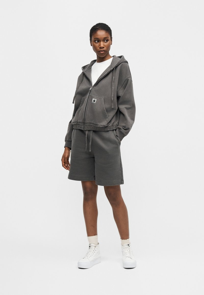 Donna che indossa felpa con cappuccio grigio scuro con cerniera e pantaloncini abbinati, maglietta bianca, calzini bianchi e sneakers alte bianche, in piedi su sfondo bianco.