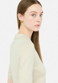 Top corto a maniche corte di un beige chiaro con una texture morbida e un colletto. Orecchini di perle in tono oro aggiungono un accento discreto.