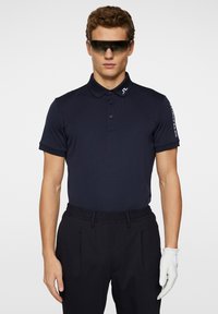 Marinblå polo skjorta med krage och tre knappar, kombinerad med mörka byxor och en vit handske. Solglasögon fulländar looken.