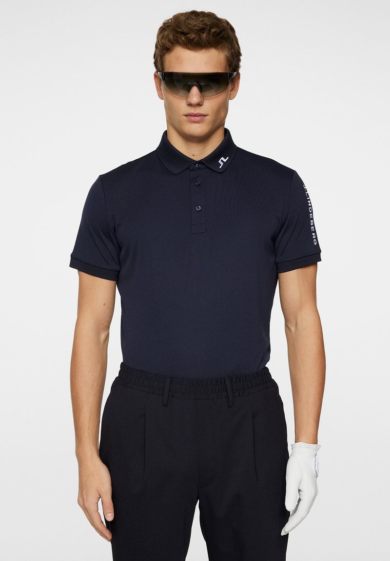 Marinblå polo skjorta med krage och tre knappar, kombinerad med mörka byxor och en vit handske. Solglasögon fulländar looken.