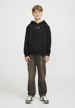 Sudadera negra con un bolsillo frontal y una etiqueta en el pecho, combinada con jeans marrones desgastados y zapatillas de deporte en blanco y negro. Diseño sencillo.