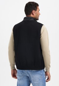 Schwarze und beige gestrickte Sweater-Weste mit hohem Kragen, strukturierten Ärmeldetails und geripptem Saum. Glattes Gewebe kontrastiert mit gestrickten Mustern.