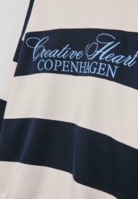 Tessuto a righe blu navy e crema con ricamo in azzurro chiaro "Creative Heart COPENHAGEN". Texture morbida, design casual e branding evidente.