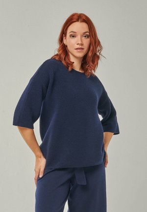 Young woman with red hair wearing a navy ribbed knit top with elbow-length sleeves and matching navy pants against a plain gray background.