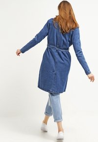 Dlouhá modrá džínsová bunda s páskem v pase, ohrnutými rukávy a rozparkem na zadní straně. Spárovaná s lehkými džínami s manžetami a bílými botami.