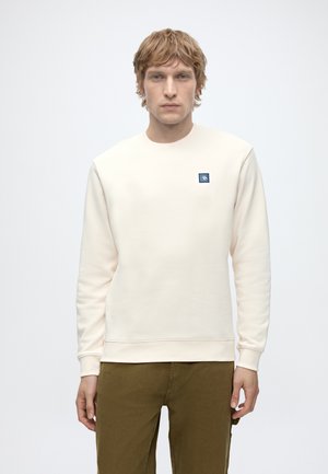 Jonge man met lichtbruin haar, die een crèmekleurige trui draagt met een klein blauw logo op de borst en olivegroene broek, staand tegen een witte achtergrond.