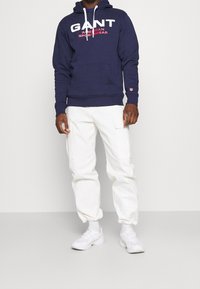 Sudadera con capucha azul marino con logo blanco "GANT AMERICAN SPORTSWEAR", bolsillo canguro, combinada con pantalones cargo blancos y zapatillas blancas.