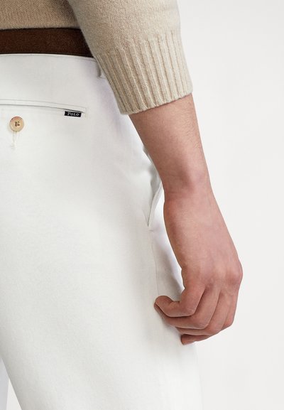 Białe bawełniane spodnie z zapięciem na guzik i boczną kieszenią. Modelka nosi beżowy sweter z prążkowanym wzorem, ukazując część brązowego paska.