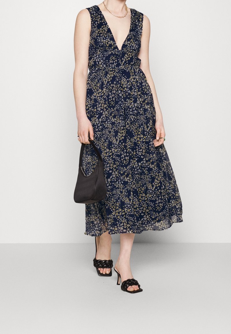 Femme portant une robe sans manches à fleurs bleu marine, tenant un sac à main noir et portant des sandales à talons noires avec des brides tressées.