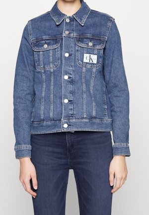 Persona che indossa una giacca di jeans blu con chiusure a bottoni e due tasche frontali con pattina sopra jeans di denim scuro.