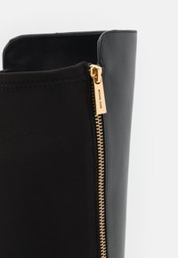 Botte en cuir noir avec une fermeture éclair dorée Michael Kors partiellement ouverte, révélant un int érieur en tissu lisse.