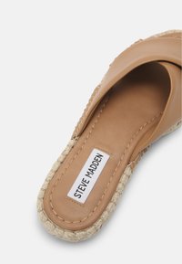 Sandale à semelle en corde tissée en cuir tan, avec une bride entrecroisée, arborant une étiquette blanche avec "Steve Madden" en texte noir.