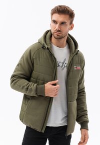 Giacca zip-up verde oliva con frontale trapuntato, materiale in pile caldo, cappuccio e polsini a coste. Indossata sopra una t-shirt grigia chiara.