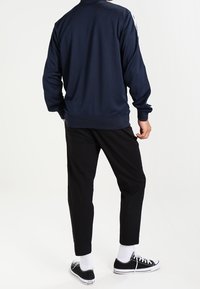 Marinblå sportjacka med långa ärmar, ribbade muddar och ett vitt rand längs axlarna, kombinerad med svarta smalrandiga byxor och svarta sneakers.